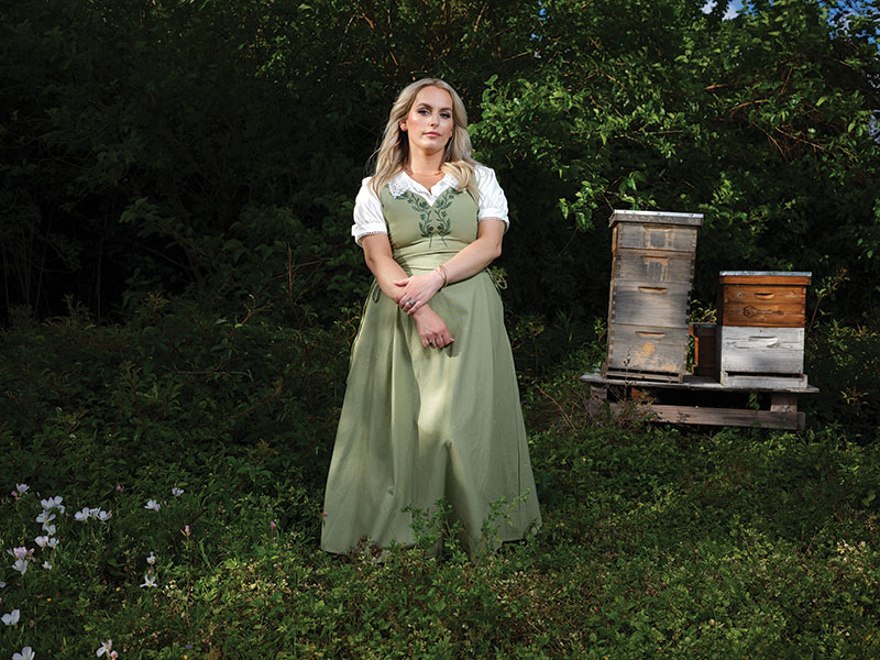 Bravender in a long green dress, in front of foliage, crates of bees (closed) behind her.