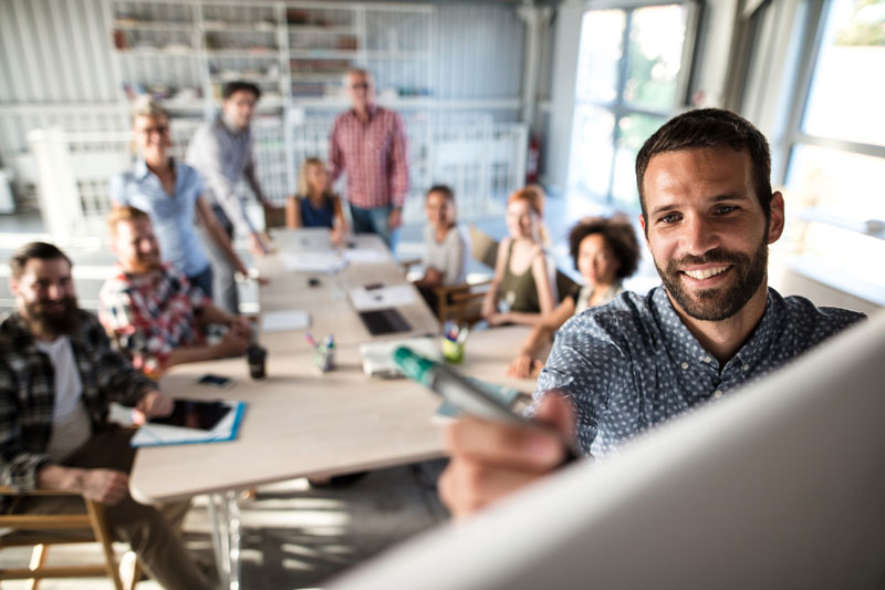 Workers in a room, focus on man at front writing on whiteboard.