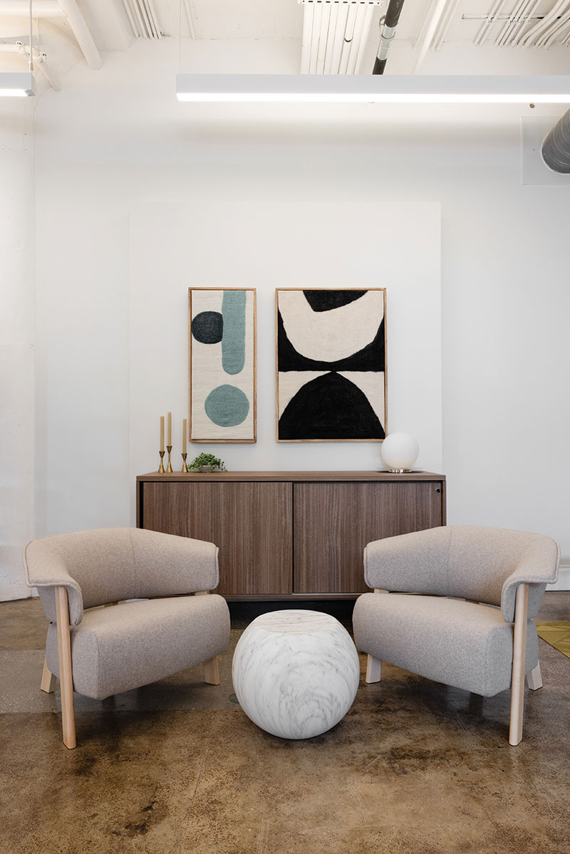 A small seating area with two beige cushioned seats and a small, almost orb-like table in between them with the top flattened. Behind the seats is a low cabinet with candles and a small spherical lamp. Above are two framed abstract art pieces.