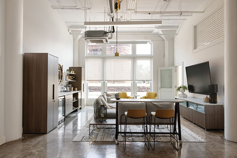Angle of photo shows KV Workspace's neighbor the Odgen Museum through window (directly facing photo angle). Room has a large TV to the right, a comfortable lounging couch seated in front of it. Behind the couch are two tall cabinets with a lower middle section in between that features drink fridges and other refreshment accessories.