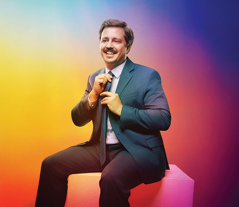 Knudsen is sitting on a low white cube-chair. He is smiling and looking to his right while adjusting his tie.