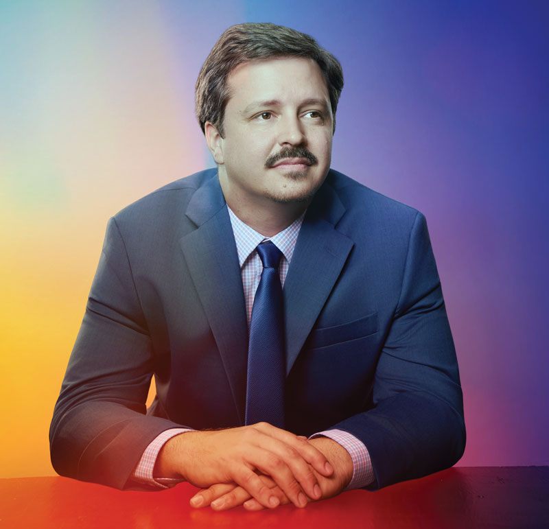 Seth Knudsen portrait. Seth is a brown-haired man with a mustache. In this series of photos he sports a gray-blue suit with a navy tie and a gray and blue small plaid button shirt underneath. In this photo he is leaning forward on a table and looking to his left, away from camera.