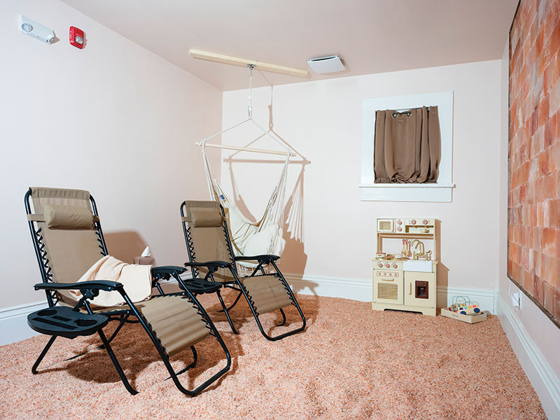 Room with pink Himalayan salt covering the floor. There are children's toys in the right corner and a hammock on the left. Towards the center are two chairs to lounge in, with one sporting a blanket.