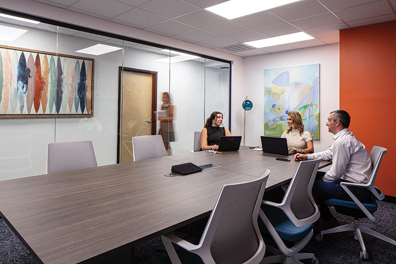 Interior of meeting room. The walls are white and orange with large artworks. The central table is a dark wood and is surrounded by gray and blue desk chairs. In this photo three people are in a meeting.