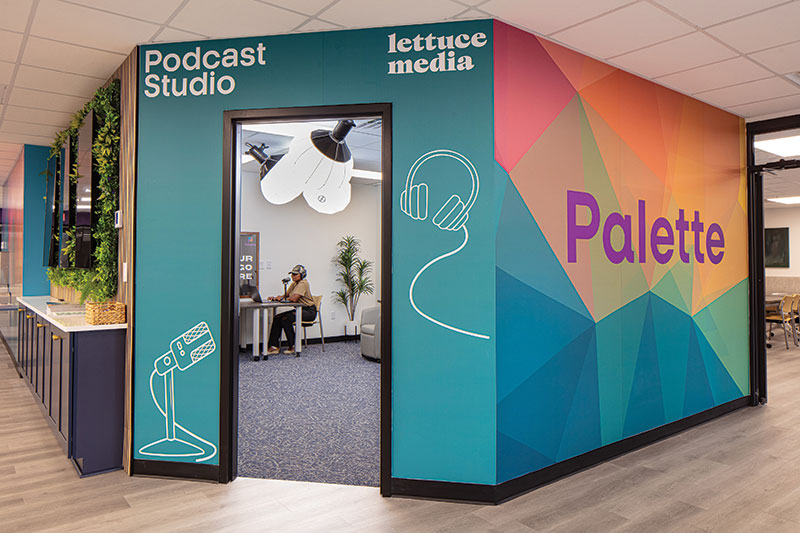 A closer look at the Podcast Studio entrance. The wall is teal with line illustrations on the left and right of the doorway. The left illustration is a mic and the right illustration is a set of headphones. In the top left around the doorway the words "Podcast Studio" can be seen. And to the right corner. "lettuce media." The right exterior wall of the Podcast Studio room is the colorful mural from before with the Palette word logo on it.