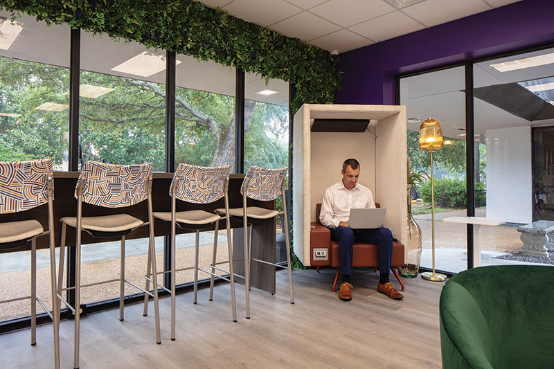 Corner of the main workspace. This corner features tall almost floor to ceiling windows. The left wall is covered with faux plants and the right wall is purple. There is a boxy chair with outlets that a man is sitting in and working in in the most corner part of this area. The to left against the windows is a tall desk/table set up with many white chairs with line patterns facing the window.