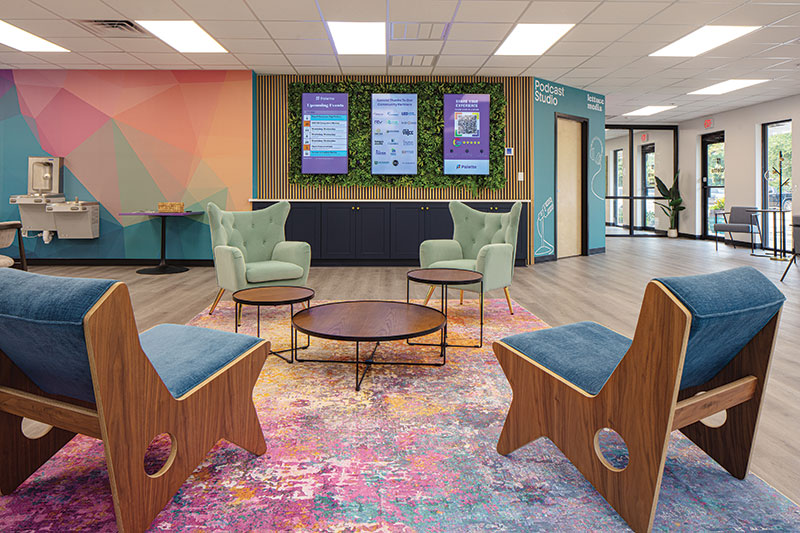Coworking Interior of Palette; the walls feature colorful but muted paints, wood paneling with faux leaves to support the TV display area. There are cozy green chairs (a pair) sitting across from more angled blue upholstered chairs with wood sides. Between them lies three round table with slightly varying heights and widths.