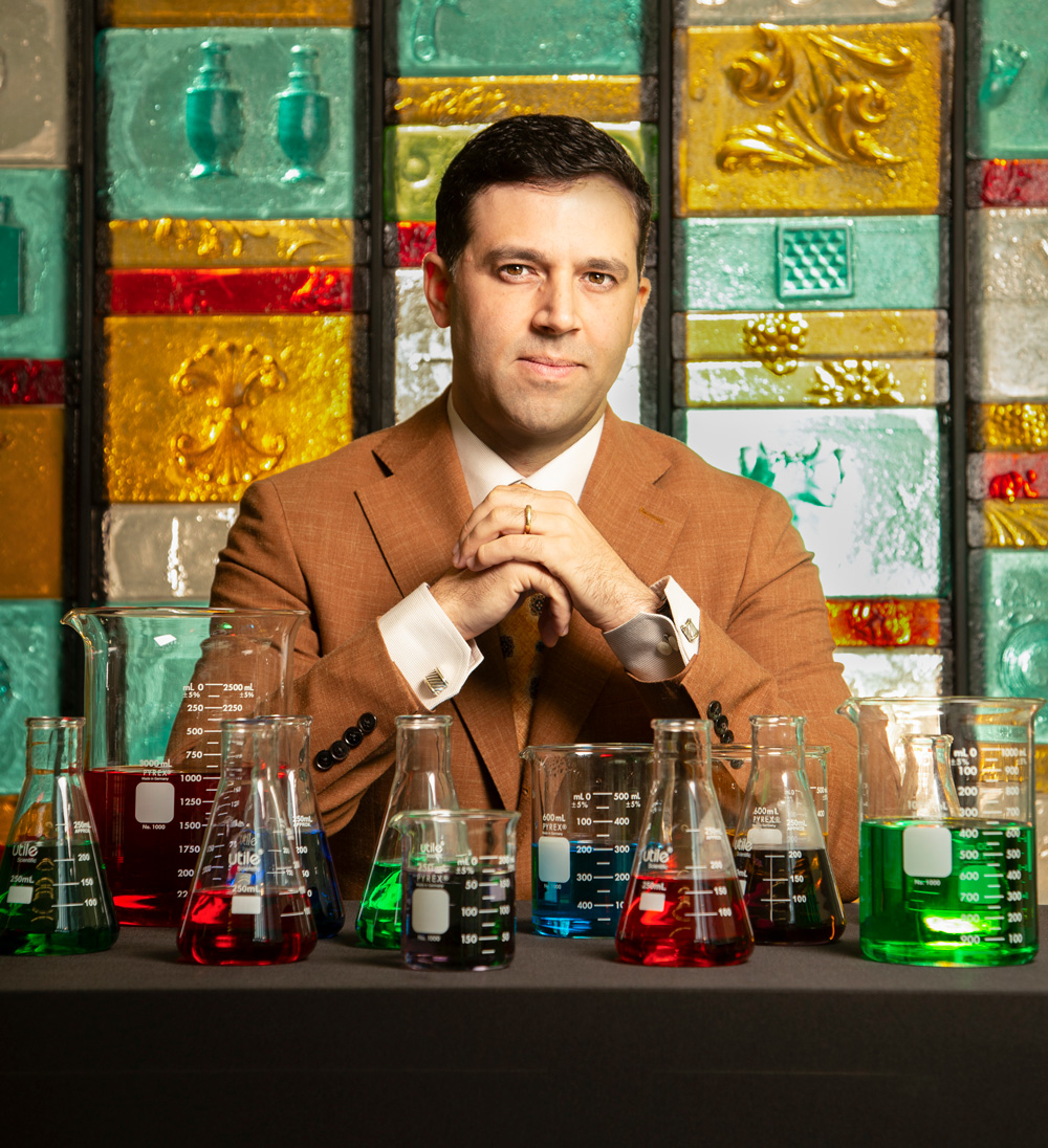 Portrait of Kris Khalil sitting with his elbows up on a table; his left hand cupping the right while he looks at the camera. He is wearing a caramel-colored suit jacket. In front of him on the table lie many flasks and beakers of varying colors. Behind him are large colored glass tiles on the wall.