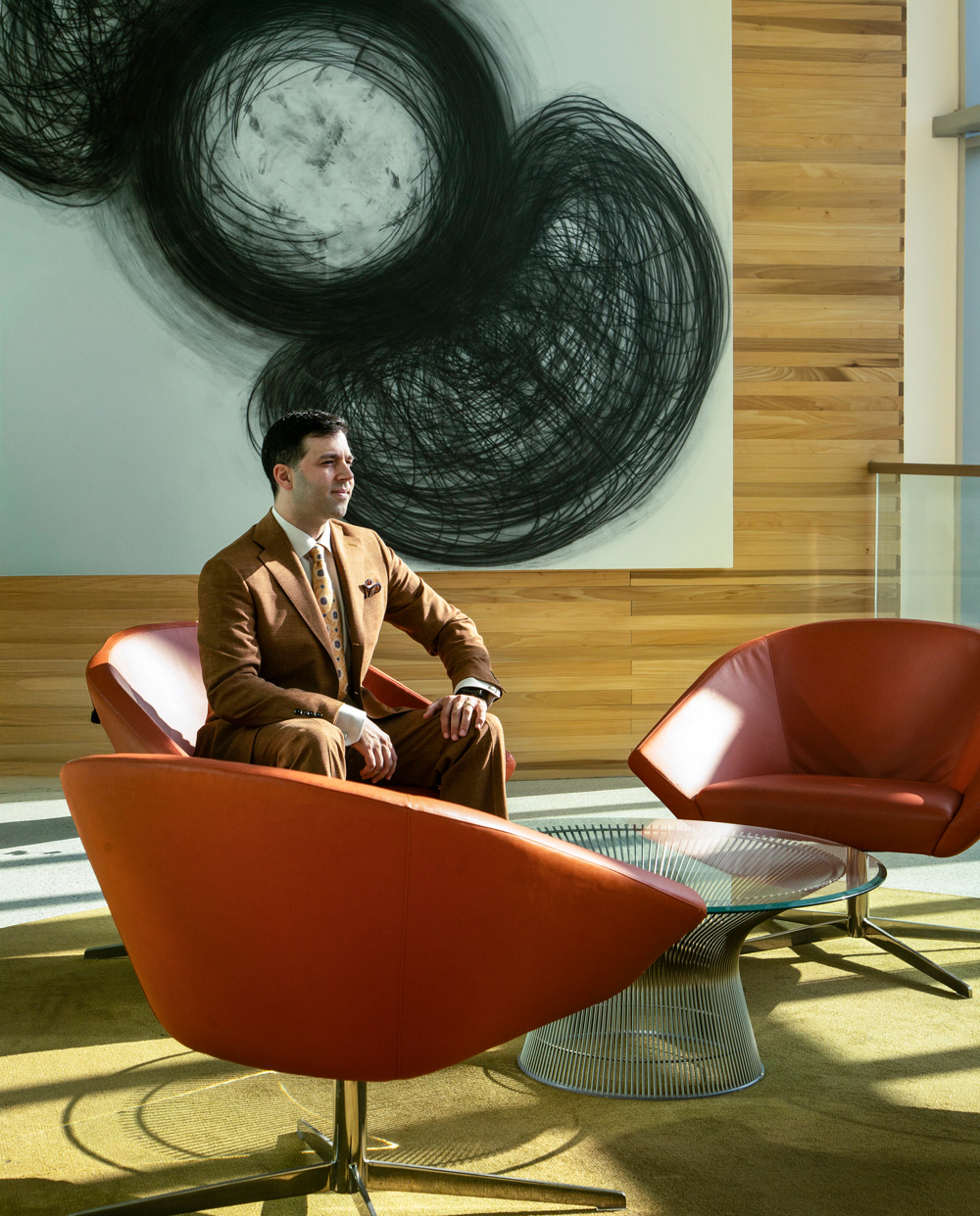Khalil seen in a curated lounge space. He is seated on a modern rust-red chair. There are two others on either side, and a glass table at the center. Behind him is an artwork in black and white.