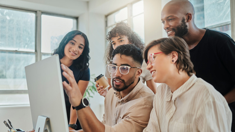 A multi-ethnic group of workers all joined behind a computer screen, looking at the same screen