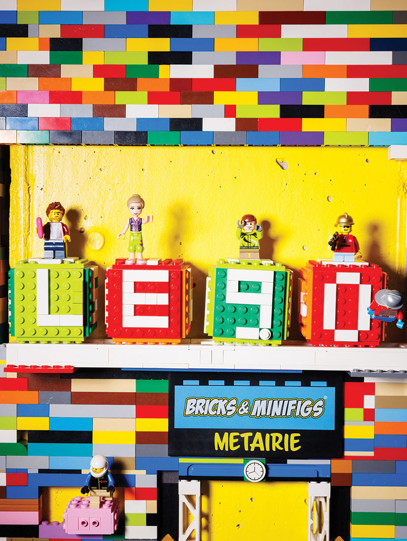 Legos arranged into 4 cube structures that with letters that spell out Lego. Each cube has a mini figure on top of it. Underneath there is a small lego-constructed Bricks & Minifigs Metairie storefront.