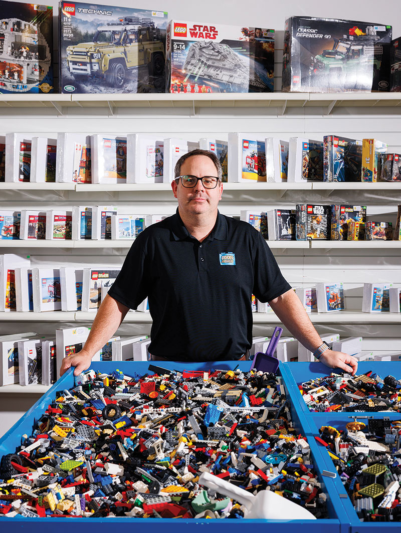 Luke Fontenot in front of three large blue bins full of Lego pieces. Behind him are repackaged Lego sets.
