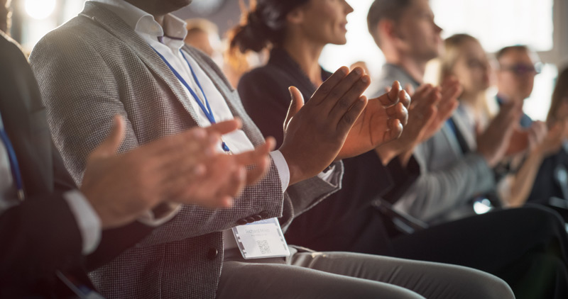 People clapping at a conference. The photo cuts off near the necks, so we only see the crowd's seated lower halves and their hands clapping