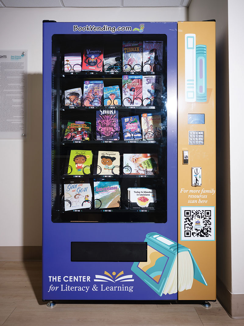 Front facing view of the book vending machine. It's main body is purple, and the panel with the keypad is yellow. There are book images on the yellow panel and near the bottom vending area. And the bottom left corner says "The Center for Literacy and Learning"