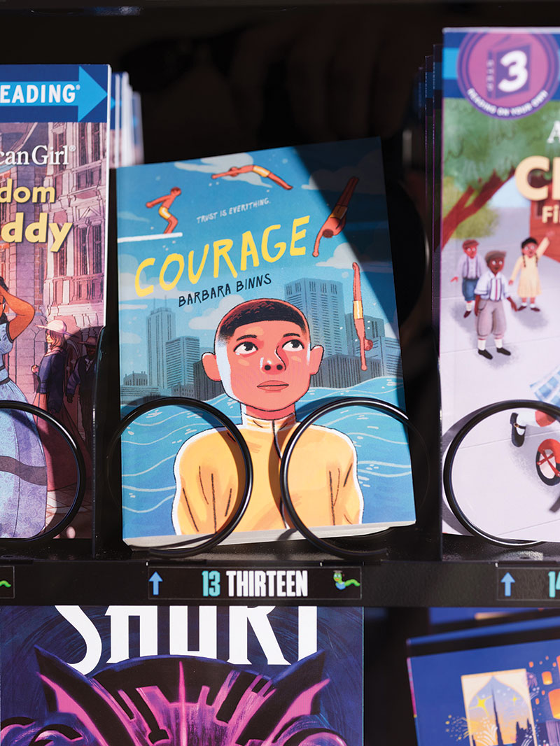 Close up of one book within the vending machine, "Courage" by Barbara Binns. The cover illustration is of a young boy front and center taking up the bottom half of the book, body facing forward while he looks above him over his left shoulder at a person diving. The top art is the various stages of diving this person is posed in