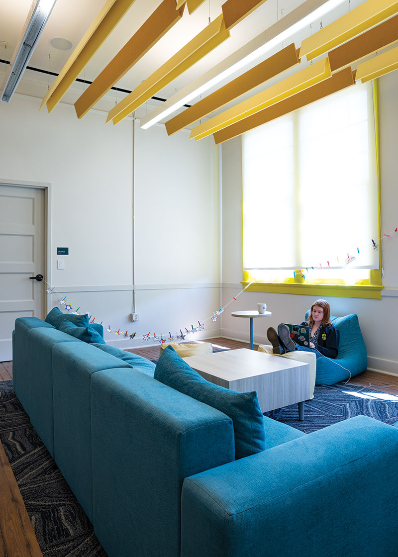 Lounge area with a wide teal couch, a coffee table, and a bean bag chair. Above are bright yellow beams